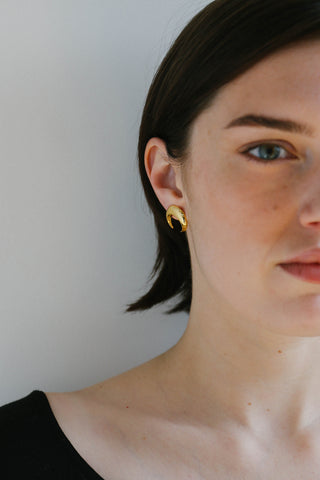 A close-up of a woman with short dark hair wearing a small gold crescent-shaped hoop earring against a plain light gray background.