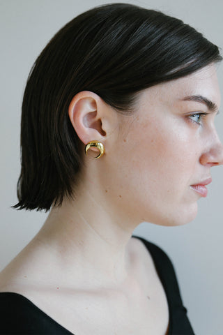 A close-up side profile of a woman with short dark hair wearing a small gold hoop earring with a crescent moon design.