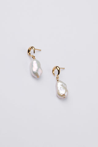 A pair of crescent moon shaped gold earrings with natural baroque pearls hanging, against a light grey background.