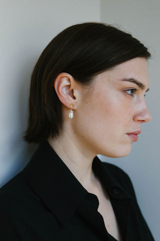 A woman in profile wearing a small gold stud earring with a white teardrop pearl drop, dressed in a black collared shirt against a neutral gray background.