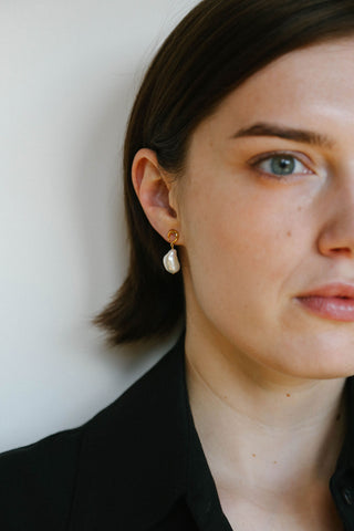 A close-up portrait of a woman wearing a delicate gold hoop earring with a small white pearl drop pendant, set against a plain light background.
