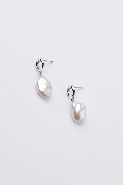 A close-up side profile of a person wearing a delicate silver hoop earring with a small white teardrop-shaped pearl drop, set against a plain light gray background.