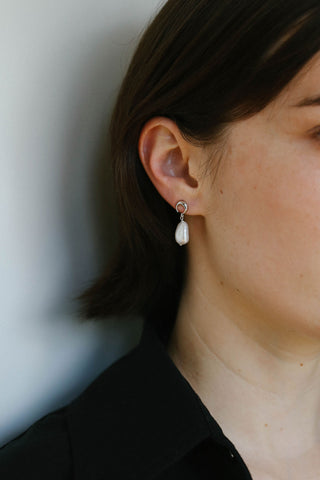 A close-up side profile of a person wearing a delicate silver hoop earring with a small white teardrop-shaped pearl drop, set against a plain light gray background.