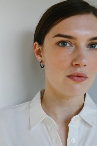 A close-up portrait of a woman wearing a black hoop earring, dressed in a white collared shirt against a plain light gray wall.