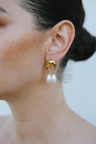 Model wears gold plated earrings by Miro Miro, crafted in a crescent moon shape from which a pair of natural freshwater tear drop pearls hang.