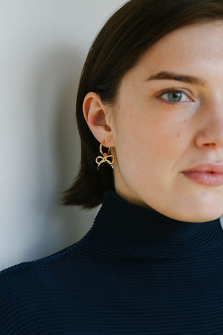 A close-up of a woman wearing a delicate gold bow-shaped hoop earring, paired with a dark navy ribbed turtleneck sweater against a neutral light gray background.