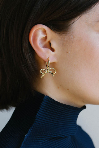 A close-up of a person's ear wearing a delicate gold bow-shaped hoop earring, paired with a dark navy ribbed turtleneck sweater.