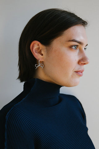 A woman with short dark hair is shown in profile wearing a silver bow-shaped hoop earring, a dark navy ribbed turtleneck sweater, and a black top layered underneath. The background is a plain light gray wall.