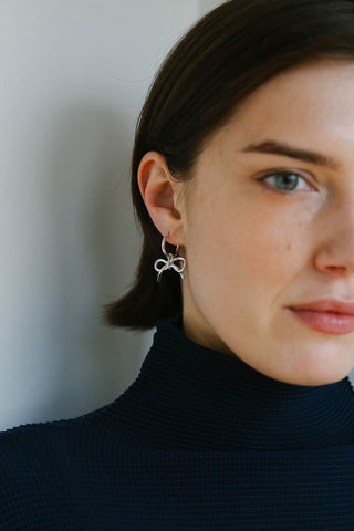 A close-up of a woman wearing a delicate silver hoop earring with a small bow-shaped charm, paired with a dark navy ribbed turtleneck sweater.