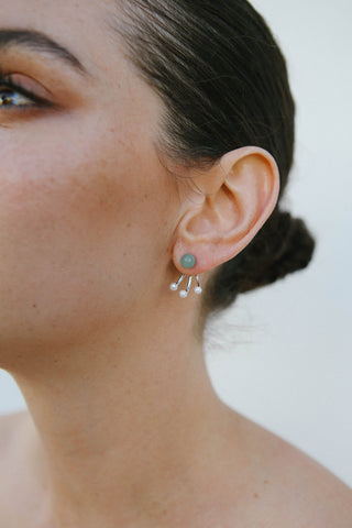Model wears rhodium plated sterling silver earrings by Miro Miro, featuring a spherical natural chrysoprase stud, with a triple curved pronged backing from which hangs small freshwater pearls.