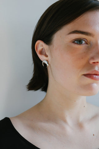 A close-up portrait of a woman with short dark hair wearing a small silver crescent moon stud earring, set against a plain light gray background.