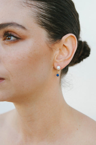 Model wears a gold plated earring by Miro Miro, featuring a freshwater pearl stud and a curved ear jacket with a spherical natural lapis lazuli stone.