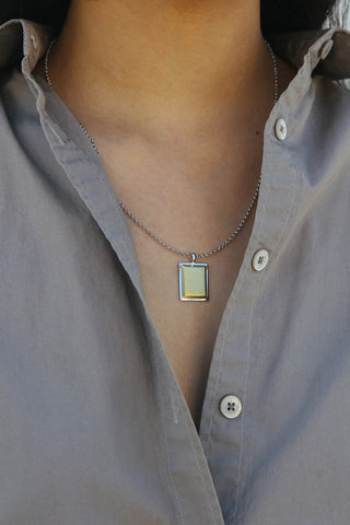 Model wears a grey button up shirt, with a necklace featuring a rectangular shape pendant, made from a two tone silver and gold finish.