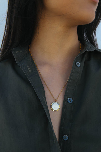 Model wears a dark green button up shirt, and a necklace featuring a circular shape pendant, made from a two tone gold and silver finish.