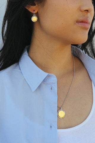 Model wears a matching earring and necklace set. The earrings feature a circular drop shape, and necklace a circular pendant, both made from a two tone silver and gold finish.